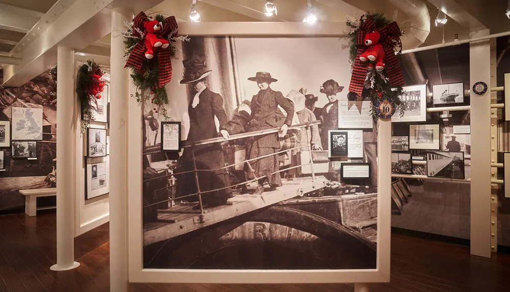 The image shows a museum exhibit featuring a large historical photograph of people boarding a ship adorned with festive decorations and red teddy bears
