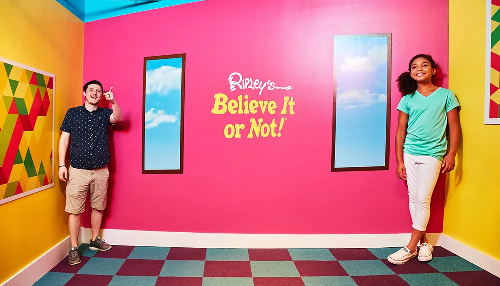 A man and a girl are standing in a colorful room with geometric patterns on the floor vibrant pink walls featuring the Ripleys Believe It or Not logo and framed images of blue skies