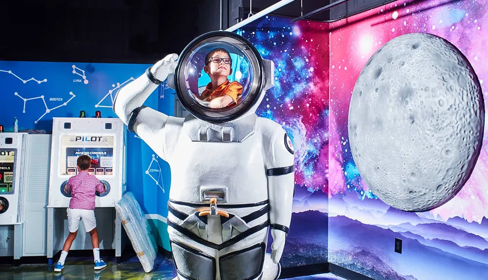 A child interacts with an interactive exhibit at a space-themed attraction while another childs face is humorously positioned in the helmet of a large astronaut statue
