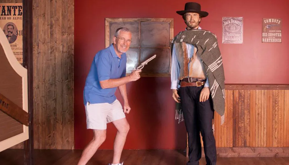 A person is playfully posing with a prop gun alongside a cowboy mannequin in a Wild West-themed setting