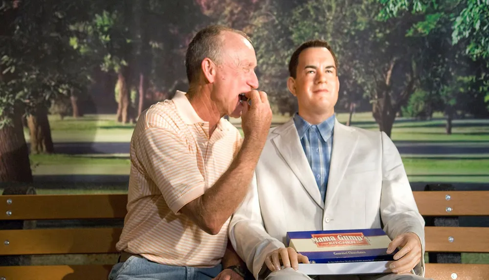 A real person is sharing a laugh next to a lifelike figure seated on a bench the latter holding a box of chocolates
