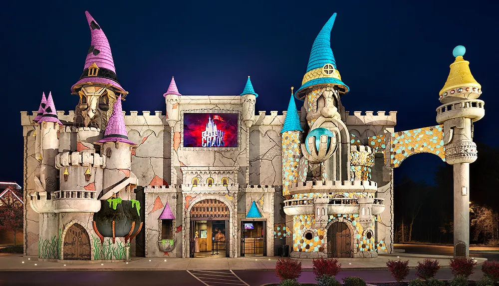 The image depicts a whimsical colorful fantasy castle facade complete with turrets and a large cauldron at night