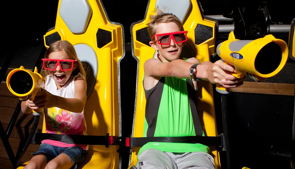 Two children wearing 3D glasses are seated in an interactive ride holding toy guns and appearing excited