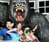 A family is posing with exaggerated expressions of fear as if they are being attacked by a large gorilla in what looks to be a staged amusement park photo opportunity