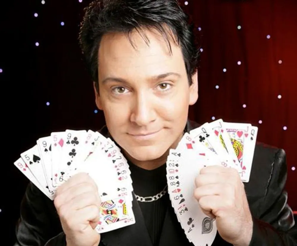 A person is posing with a fan of playing cards in both hands wearing a black outfit and a necklace with a backdrop featuring twinkling lights