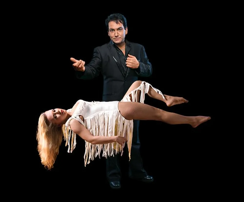 A man appears to be magically levitating a woman horizontally in front of him against a black backdrop