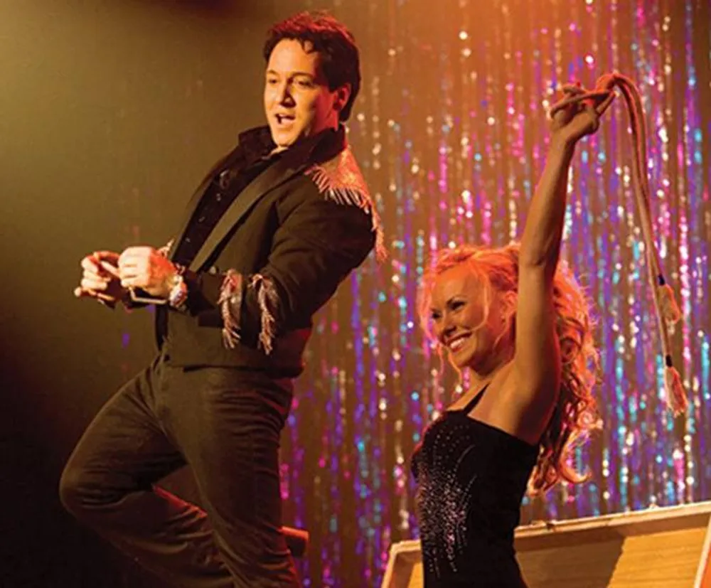 A man and a woman are energetically performing a dance routine on stage with the man mid-leap and the woman holding a prop against a sparkly backdrop