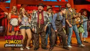 The image shows a lively group of performers on stage, likely from a Hatfield & McCoy-themed dinner show, with some playing musical instruments and others singing or acting, evoking a sense of fun and entertainment.