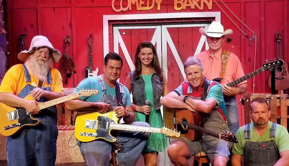 A group of six performers some holding guitars and one with a microphone are posing on a stage decorated to resemble a barn with a sign reading Comedy Barn in the background