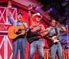 A group of six performers some holding guitars and one with a microphone are posing on a stage decorated to resemble a barn with a sign reading Comedy Barn in the background