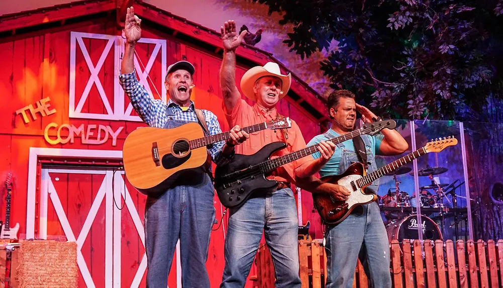 Three performers on a stage with musical instruments are engaging with an audience in front of a barn-like backdrop labeled THE COMEDY