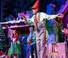 A festive Christmas performance is taking place on stage featuring lively musicians a decorated tree and a performer dressed in holiday-themed attire