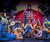 A lively group of people in colorful outfits including overalls and gingham dresses perform energetically on stage with a monkey puppet amidst a festive setting