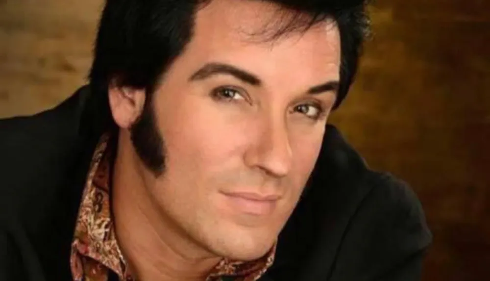 The image shows a person with dark sideburns and styled black hair wearing a black jacket and a patterned shirt or scarf looking towards the camera with a slight smile against a blurred brown background