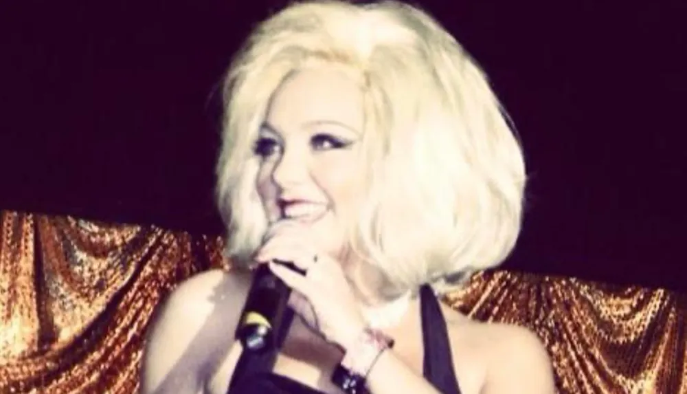 The image shows a person with platinum blonde hair singing into a microphone with a golden background giving the impression of a live performance or event