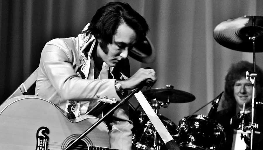 A performer dressed in an Elvis Presley-style outfit is intently playing a guitar on stage with a drummer in the background.