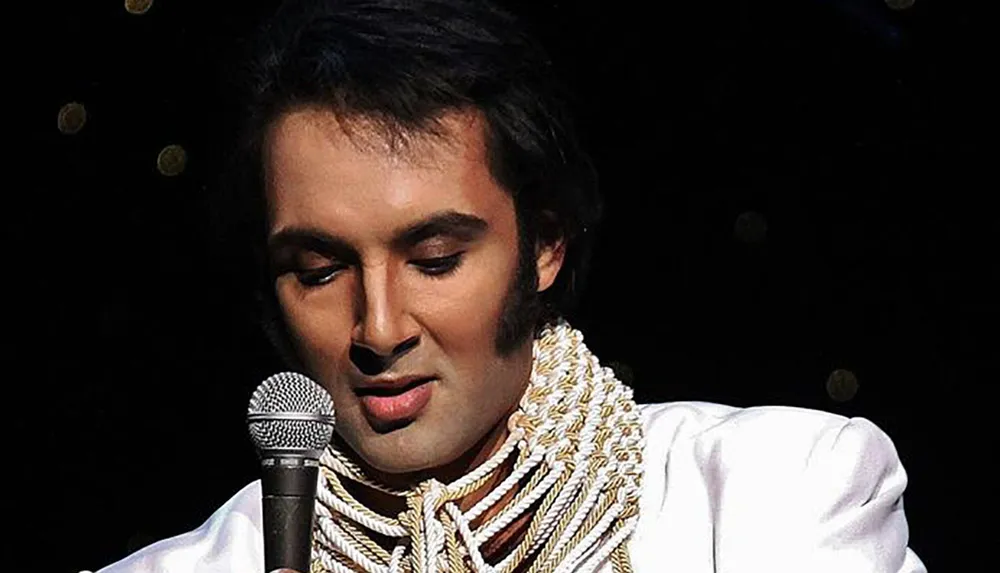 A performer wearing an elaborately decorated white jumpsuit and cape reminiscent of Elvis Presleys iconic stage costumes is singing into a microphone