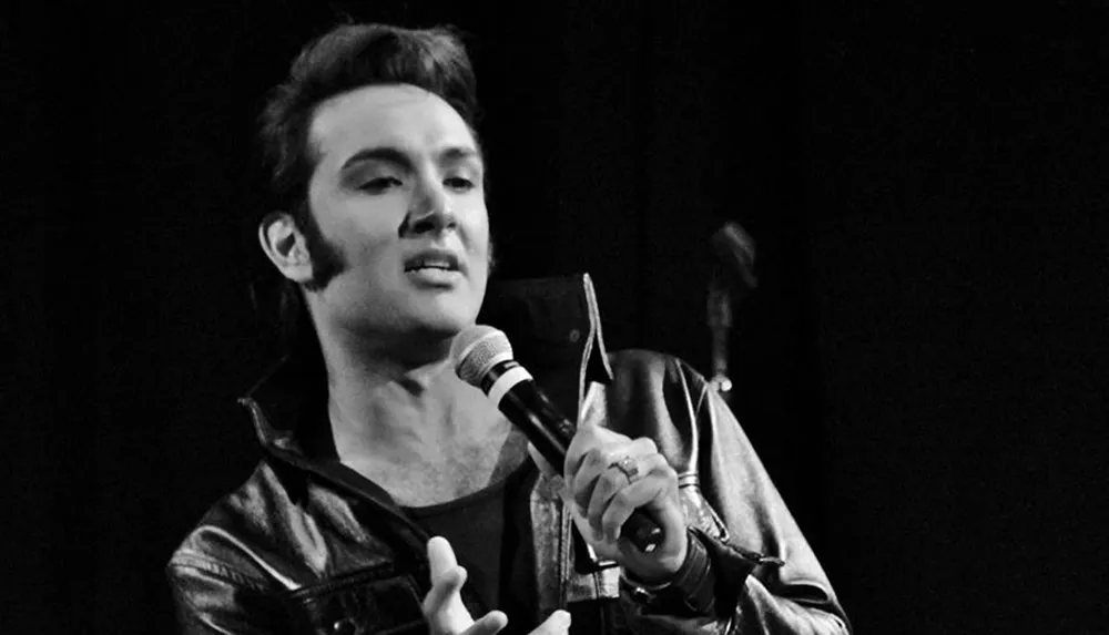 A person wearing a leather jacket is singing into a microphone on stage in a black and white photograph