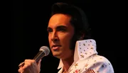 A person wearing an Elvis Presley-style costume is holding a microphone, presumably performing or about to perform.