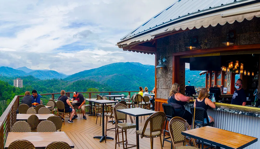 People are enjoying food and drinks at an outdoor mountain-view bar with lush green hills in the background