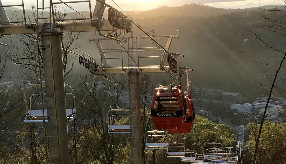 A cable car emblazoned with the name Anakeesta ascends a mountain amidst sunlit misty scenery above what appears to be a series of empty chairlifts