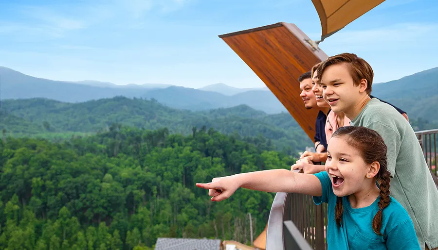 A group of people are joyfully observing a lush green mountain landscape from a balcony.