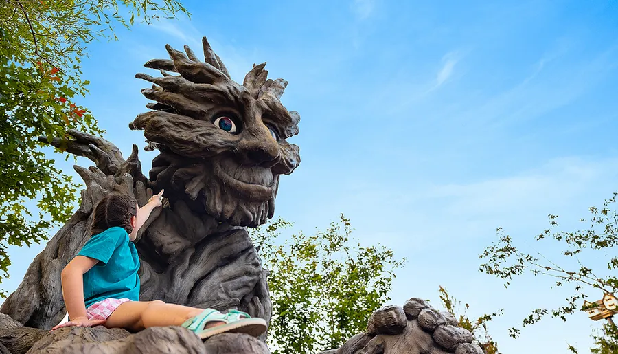 A child in a blue shirt is pointing at a large, whimsical tree-like statue with a face against a clear blue sky.