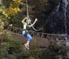 A smiling child wearing a helmet and harness joyfully zip-lines against a backdrop of trees and a waterfall