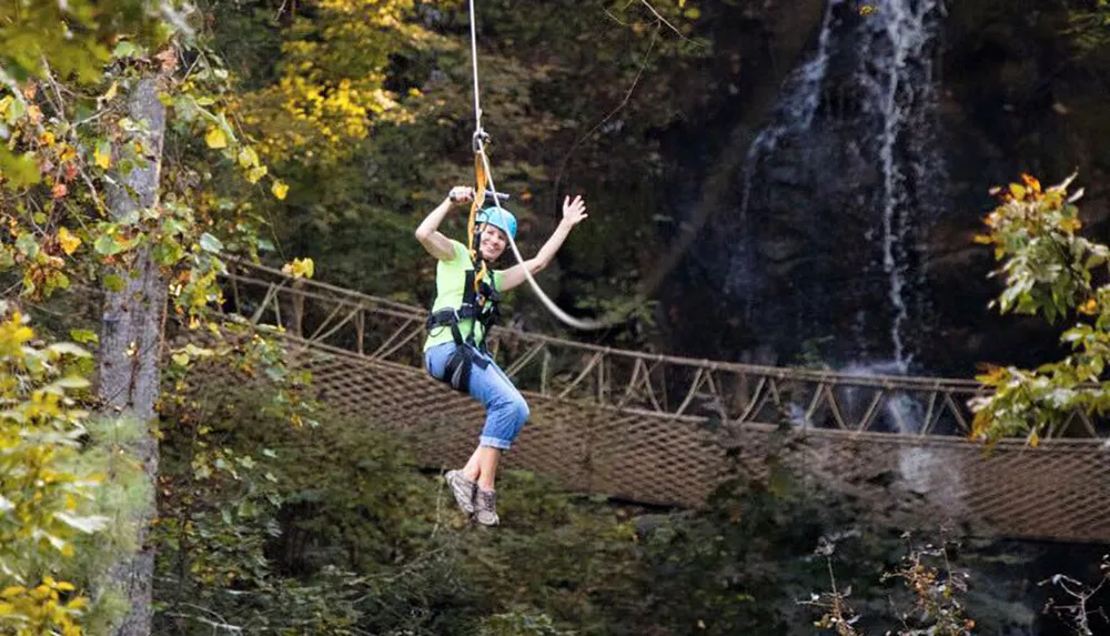A person is gleefully ziplining in a forested area with a waterfall and a suspension bridge in the background