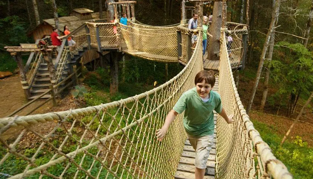 A child is smiling as he walks across a rope bridge connected to tree houses in a lush forested area while other people enjoy the treetop structures in the background