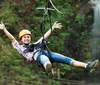 A smiling child wearing a helmet and harness joyfully zip-lines against a backdrop of trees and a waterfall