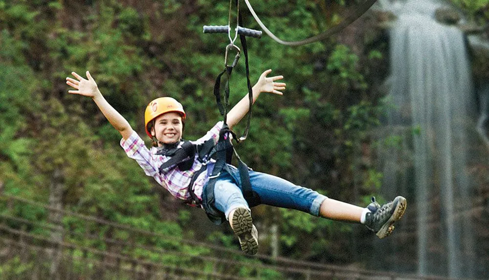 A smiling child wearing a helmet and harness joyfully zip-lines against a backdrop of trees and a waterfall