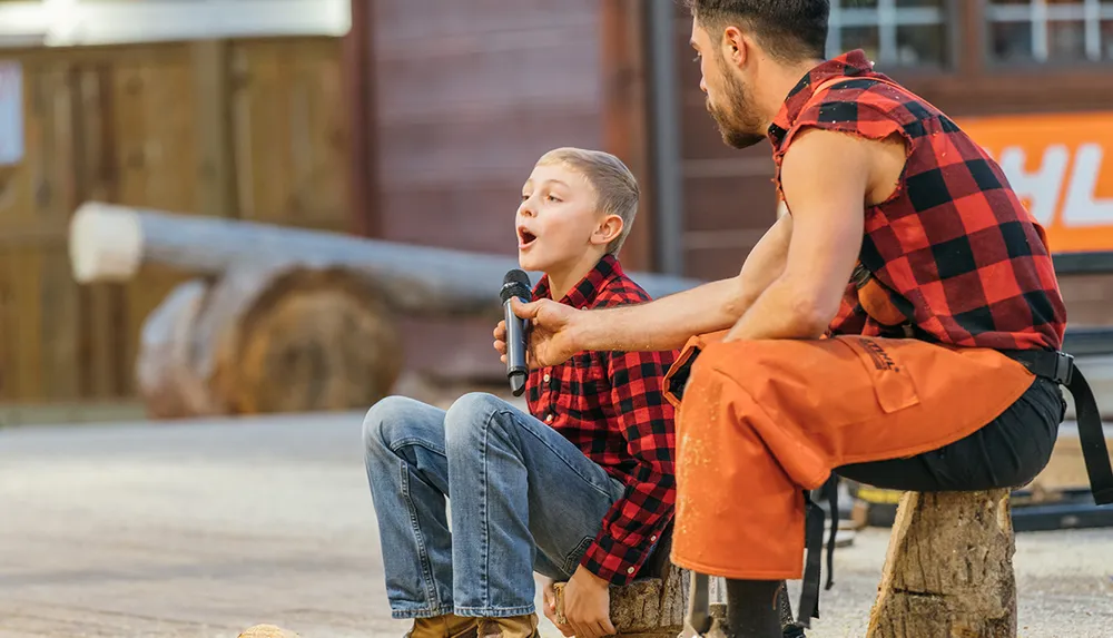 A young child in a checkered shirt appears to be singing or speaking into a microphone while sitting on a log accompanied by a person in similar attire sitting nearby with a pensive expression