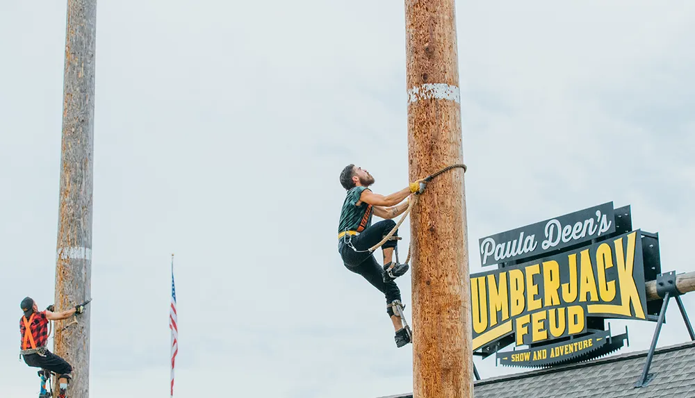 Two individuals are climbing tall wooden poles at an outdoor lumberjack event with an American flag and signage for Paula Deens Lumberjack Feud Show and Adventure in the background
