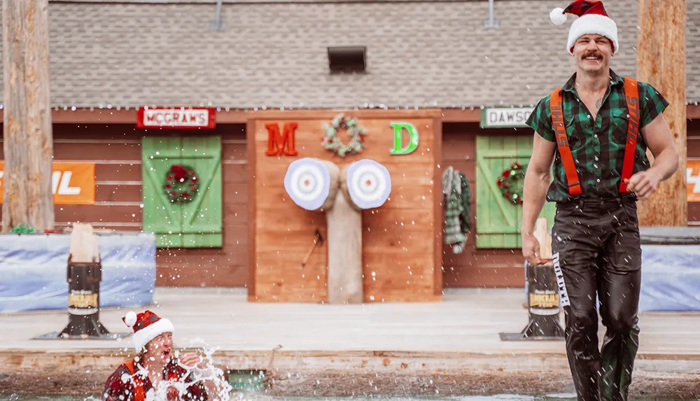 A man in a Santa hat is walking away while another in similar attire splashes in the water during a festive event with a rustic backdrop