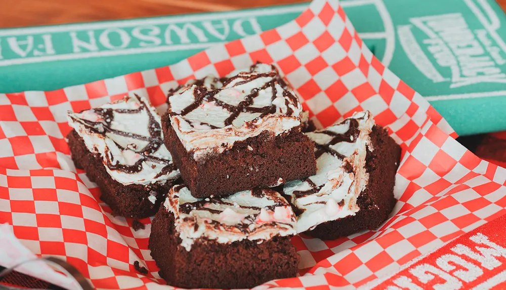A basket lined with red and white checkered paper holds chocolate brownies topped with white icing and chocolate drizzle