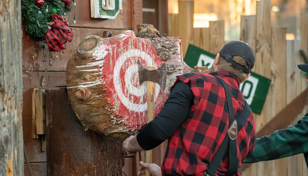 A person wearing a red and black checkered shirt is throwing an axe at a wooden target painted with a red bullseye