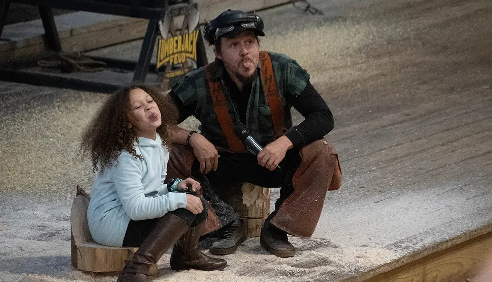 A child and an adult wearing lumberjack attire are playfully sticking out their tongues while sitting on tree stumps
