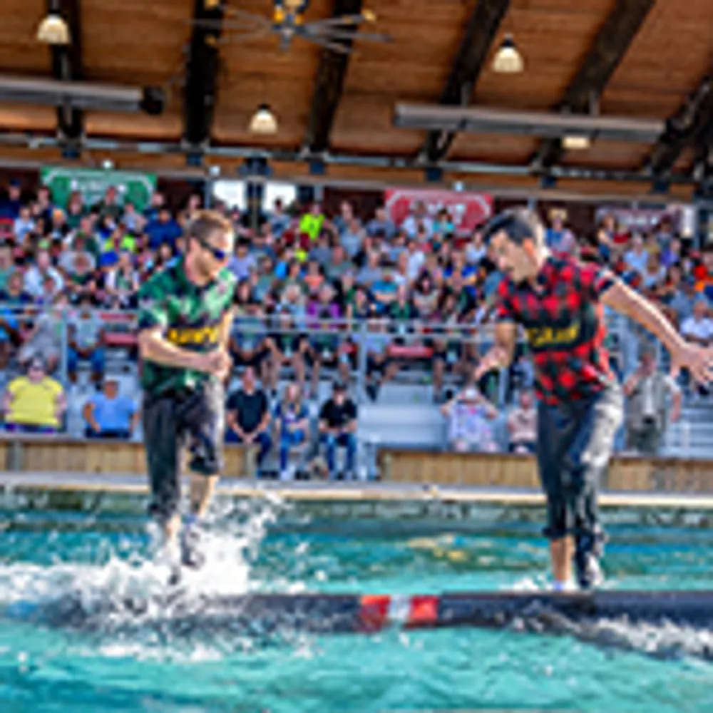 Two people are competing in a log-rolling event over water while an audience watches from the stands