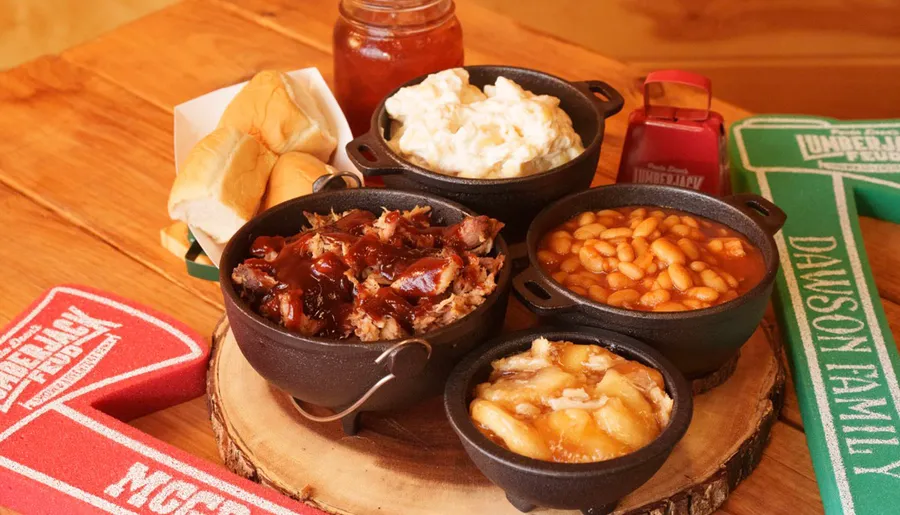 The image shows a rustic barbecue meal with pulled pork, baked beans, potato salad, and dinner rolls served in cast iron skillets and jars on a wooden table.
