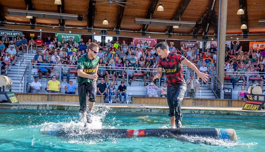 Two competitors in plaid shirts are engaging in a log rolling contest over a pool of water in front of an entertained audience.