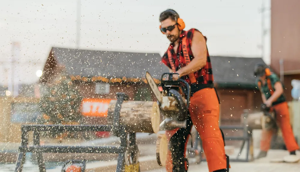 A person in safety gear is using a chainsaw to cut wood creating a spray of sawdust with another individual working in the background