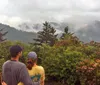 Two people are standing together overlooking a misty mountain landscape surrounded by green foliage