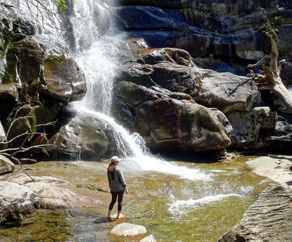 A person stands on a rock observing a picturesque waterfall cascading down rocky terrain on a sunny day