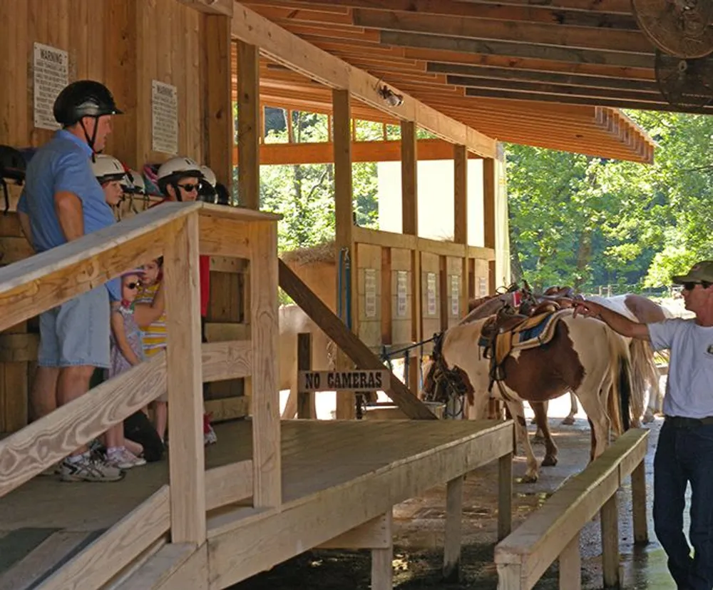 A group of people wearing helmets wait in line at a pony ride station while an attendant stands next to a pony under a wooden structure with a sign saying NO CAMERAS