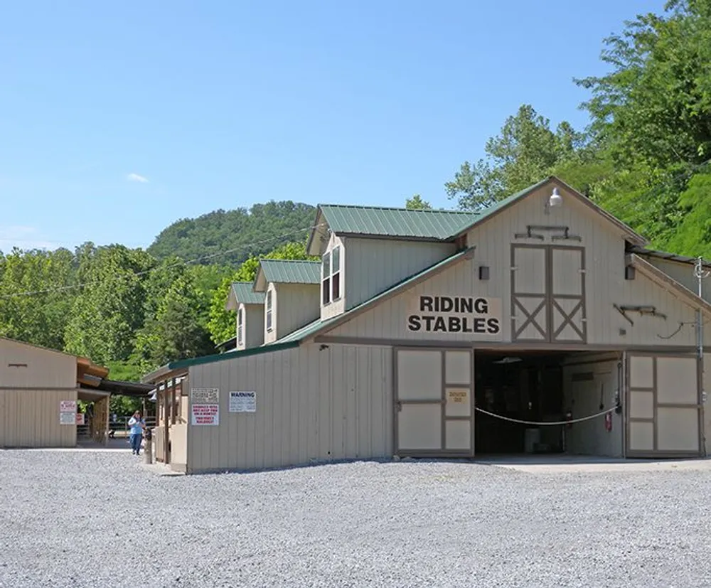 The image shows a large building with RIDING STABLES signage indicating it is a facility for horseback riding with a few people nearby set against a backdrop of lush green hills