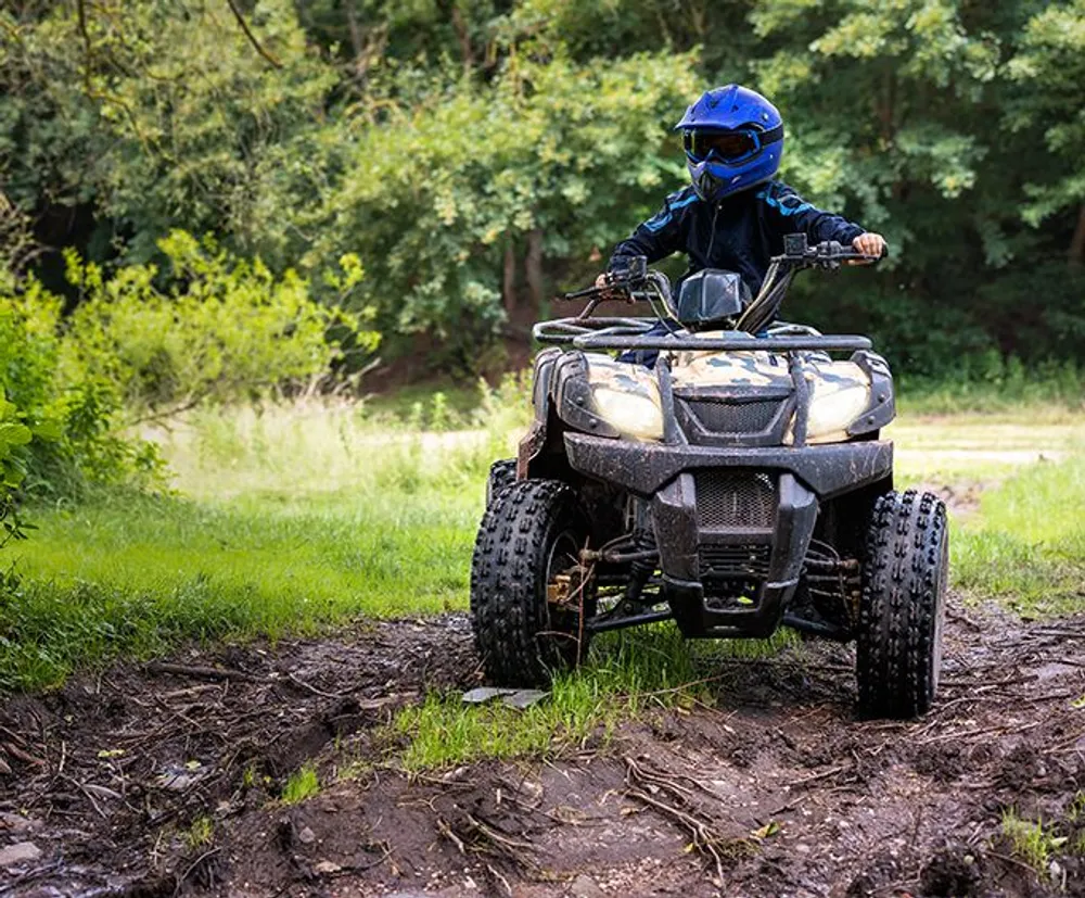 A person wearing a helmet and protective gear is riding an all-terrain vehicle through a muddy forest trail
