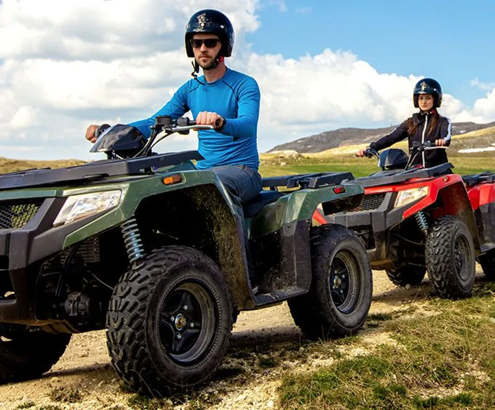 Two people are riding all-terrain vehicles ATVs across a grassy hilly landscape wearing helmets for safety