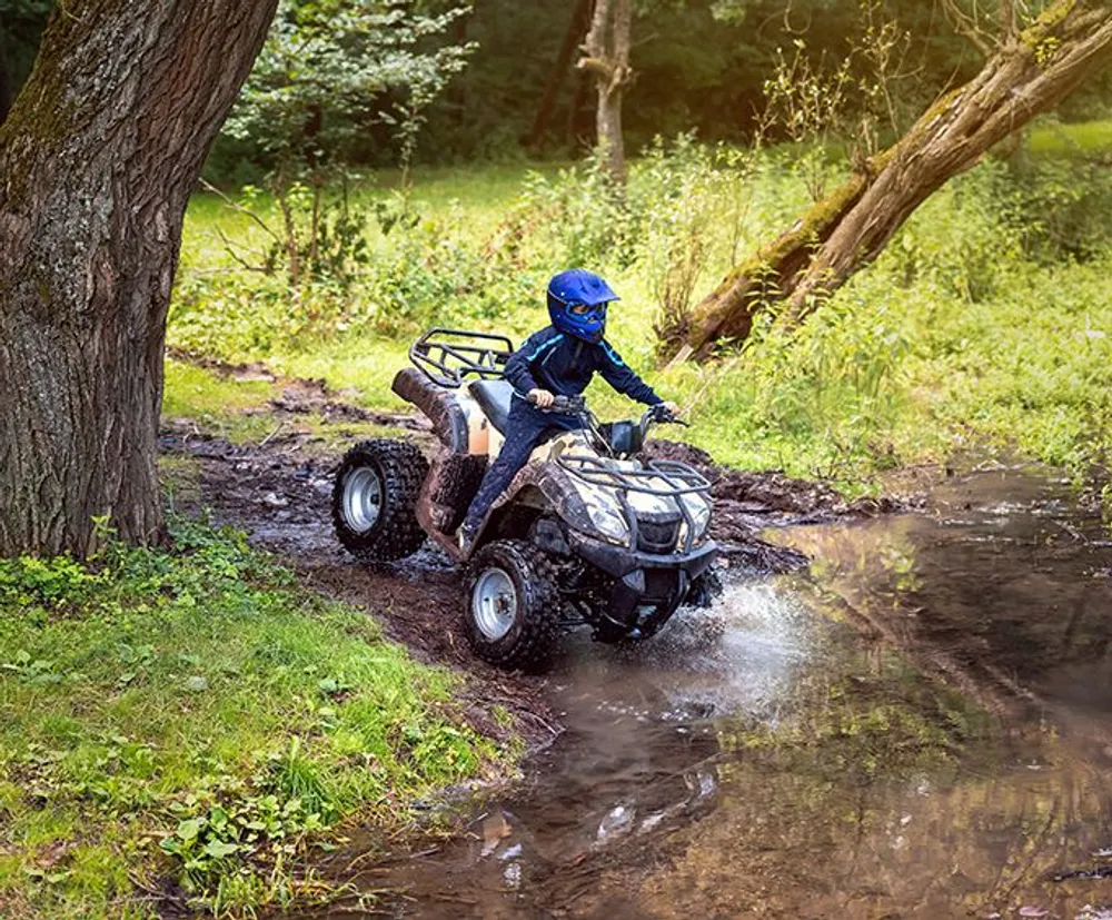 A person wearing a blue helmet rides an ATV through a muddy water puddle in a forested area