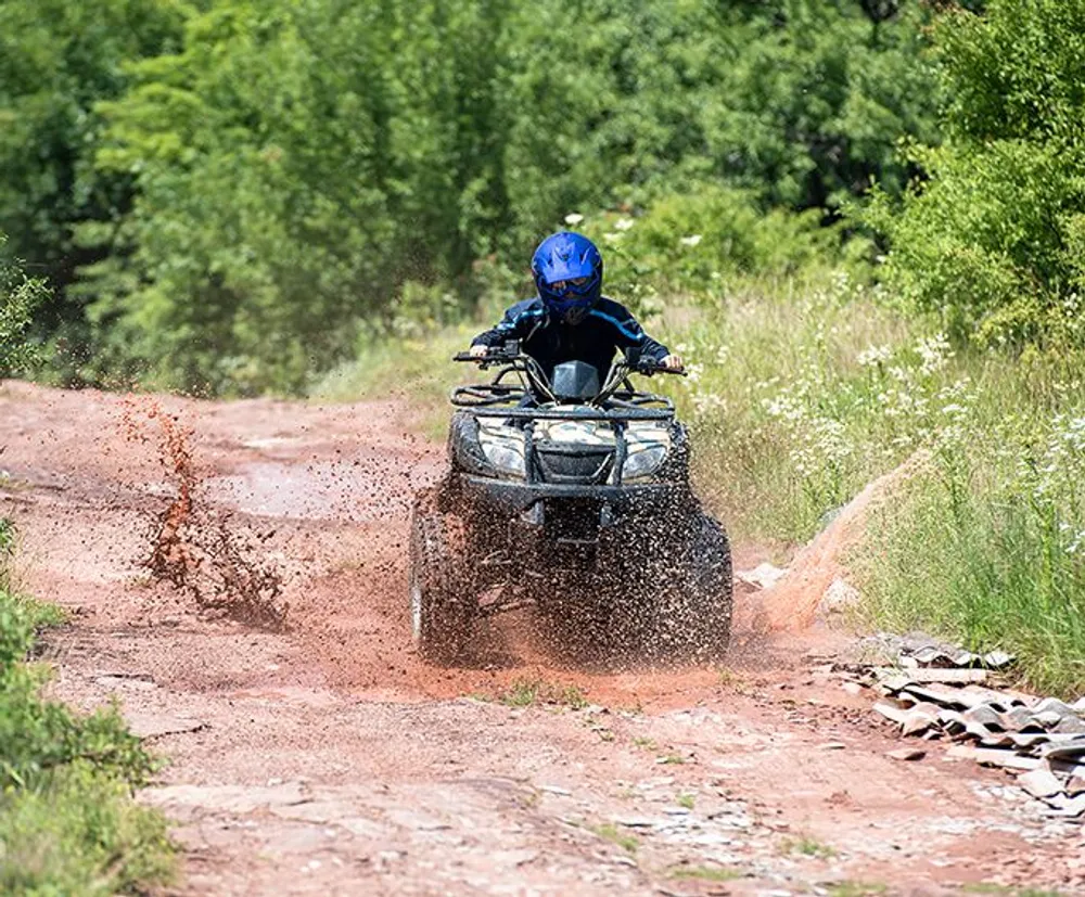 A person in a blue helmet is riding an ATV through a muddy trail splashing dirt around